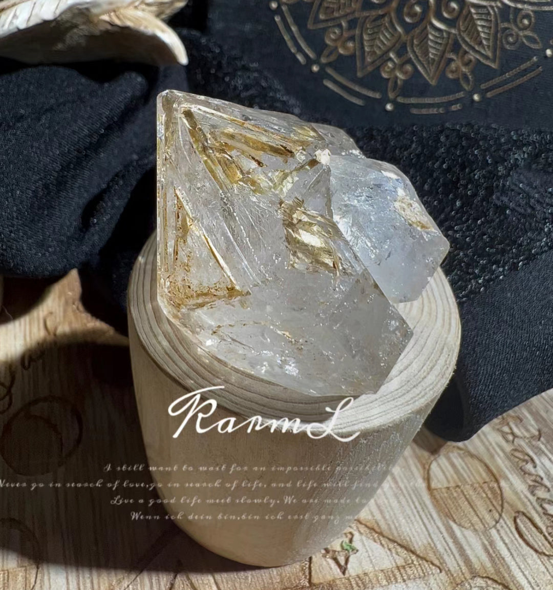 A rough quartz crystal with golden inclusions rests on an engraved stone base against a patterned textile background